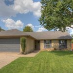 Single-story brick house with a gray roof and two-car garage, a large green lawn, a tall tree on the right, and a paved driveway under a partly cloudy sky.