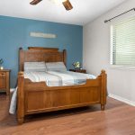 A neatly made wooden bed with striped bedding sits against a blue accent wall. A nightstand with plants and a small vase is beside the bed. The room has hardwood floors and a large window with blinds.