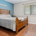 A cozy bedroom with a wooden bed frame, blue accent wall, striped bedding, wooden nightstand with a plant and lamp, and a window with blinds letting in natural light onto wood flooring.