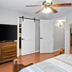 A bedroom with wood flooring, a bed with striped bedding, a wooden dresser with a large TV, double white doors, a ceiling fan, a tall mirror, and a shelf with small decor items on a gray wall.