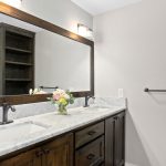 A bathroom with a double sink vanity, marble countertop, two black faucets, a large framed mirror, wall-mounted light fixtures, a towel bar, dark wood cabinetry, and a vase of flowers on the counter.