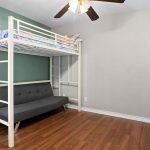 A room with a white metal loft bed, a colorful mattress on top, and a dark gray futon below. The room has light gray and green walls, wood flooring, a white door, and a ceiling fan with lights.