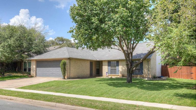 A single-story brick house with a gray roof and attached two-car garage, surrounded by a well-kept lawn and trees, sits on a quiet residential street under a partly cloudy sky.