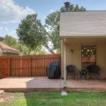 A backyard patio with stamped concrete, a wooden fence, a black grill, two chairs with a small table under a covered area, and a fire pit on the grass. The sky is partly cloudy.
