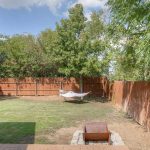 A backyard with a grassy lawn, wooden fence, leafy trees, a white hammock in the center, and a small fire pit near a tiled patio area.