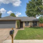 Single-story brick house with a gray shingle roof, two-car garage, and large front lawn. A black mailbox stands near the driveway, and a large tree provides shade in the yard. The sky is partly cloudy.