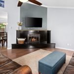 A cozy living room with brown leather sofas, a blue ottoman, a fireplace with a TV above it, hardwood floors, and a dining area visible through an open doorway.