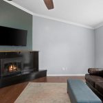 Modern living room with a dark corner fireplace, mounted TV, brown leather sofa with patterned pillows, blue ottoman, and a window with blinds. A basket and vase with flowers sit on the fireplace ledge.