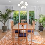 A bright dining room with a wooden table and six chairs on an orange patterned rug, large potted plants, a modern chandelier, and glass doors opening to a green outdoor view.