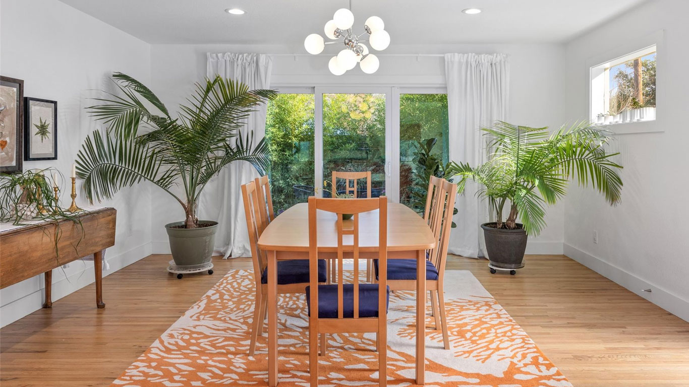 A bright dining room with a wooden table and six chairs on an orange patterned rug, large potted plants, a modern chandelier, and glass doors opening to a green outdoor view.