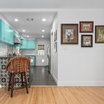 A modern kitchen with turquoise cabinets, a patterned tile breakfast bar with two wicker stools, white brick backsplash, and framed artwork on the adjacent white wall. Light wood flooring throughout the space.