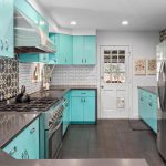 Modern kitchen with turquoise cabinets, stainless steel appliances, white subway tile backsplash, patterned accent tiles, dark countertops, and dark wood floors. A white back door with glass panes lets in natural light.