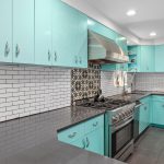 Modern kitchen with glossy turquoise cabinets, gray countertops, stainless steel appliances, white subway tile backsplash, and patterned tile above the stove. A glass door lets in natural light from outside.