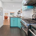 A modern kitchen with turquoise cabinets, black flower-patterned backsplash, stainless steel appliances, and a black kettle on the stove. The kitchen opens to a bright living area with wood flooring.