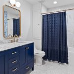 Modern bathroom with a double sink vanity featuring navy cabinets, gold hardware, and marble countertop. A white toilet, bathtub with a navy shower curtain, and blue patterned accent wall are also visible.