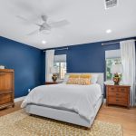A modern bedroom with dark blue walls, white ceiling, and wooden floor. The room has a bed with white bedding and yellow accent pillows, wooden nightstands, a dresser, white curtains, and a patterned yellow rug.