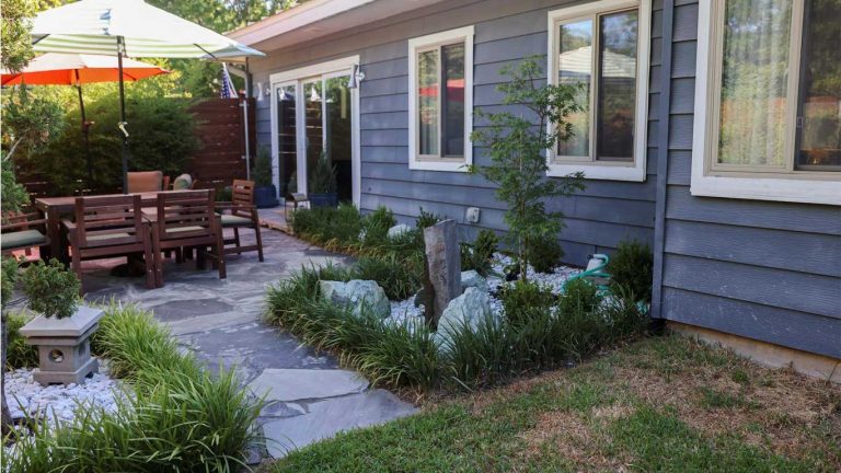 A backyard with a stone patio, wooden outdoor dining set, umbrellas, and a small landscaped garden with stones and greenery next to a gray house with multiple windows.