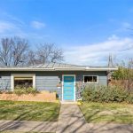 A single-story gray house with a bright blue front door, large front window, and a small brick planter filled with plants. The yard has green grass, shrubs, and leafless trees under a blue sky.