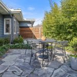 A patio with stone flooring, a round metal table and chairs, potted plants, and green bushes beside a gray house with large windows on a sunny day.