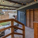 Covered patio with string lights, wooden railing and privacy slats, overlooking a backyard with lawn chairs, a small fire pit, a greenhouse, and a shed enclosed by a wooden fence.