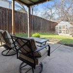 Two cushioned patio chairs and a glass table sit under a covered area in a backyard with green grass, a wooden fence, and a small glass greenhouse in the corner.