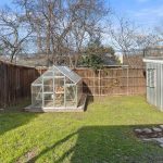 A small backyard with green grass, a wooden fence, a glass greenhouse, a metal shed, and a stone path leading to a small concrete area near the shed. Leafless trees are visible beyond the fence.