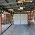 A partially enclosed garage with a white overhead door, exposed wood beams, and slatted wood fencing. The concrete floor shows stains and debris, and the walls have visible wear and damage.
