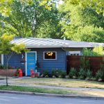 A small blue house with a bright blue door, a metal roof, and a neatly landscaped yard with shrubs, a small tree, and two red sculptures near the entrance. A wooden fence encloses part of the property.