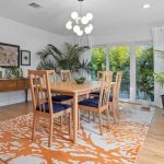 A modern dining room with a wooden table and six chairs on an orange and white patterned rug, surrounded by potted plants, framed art on the wall, and large windows letting in natural light.