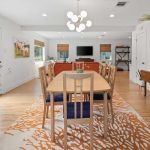 Bright dining room with a wooden table and six chairs on an orange and white rug, open to a living area with a sofa, TV, wall art, and large windows. Green plants decorate the modern, airy space.