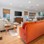 A bright living room with hardwood floors, an orange sofa, patterned armchair, wooden coffee table, TV on a wooden dresser, and large windows with bamboo shades. The open space leads to a kitchen in the background.