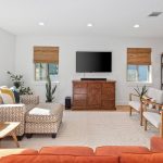 A bright living room with a red-orange sofa, patterned armchair, wooden coffee table, TV on a wooden cabinet, four white chairs, plants, and shelves; sunlight streams through windows with bamboo shades.