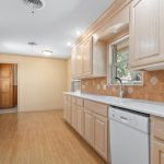 A spacious kitchen with light wood cabinets, white countertops, a white stove, a dishwasher, and tan tile backsplash. Wood flooring and warm yellow walls create a bright and inviting atmosphere.