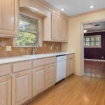 A kitchen with light wood cabinets, white countertops, tan tile backsplash, dishwasher, and window above the sink. The floor is wood, and an open doorway leads to a room with purple walls and wood flooring.