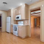 A spacious kitchen with light wood cabinets, white appliances, wood flooring, and neutral walls. The kitchen opens to two rooms, one with dark wood paneling and another with a dark accent wall. A window lets in natural light.