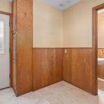 A small room with wood-paneled walls, a white door with a window, and a doorway leading to a bathroom with a white sink and towel bar. The space has light-colored flooring and cream upper walls.