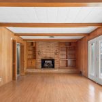 A living room with wood-paneled walls, wood floors, exposed ceiling beams, a brick fireplace flanked by built-in shelves, and double glass doors leading outside.