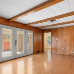 A bright, empty room with wood-paneled walls, light wood flooring, exposed ceiling beams, and three glass doors leading outside. Electrical outlets and a vent are visible on the back wall.