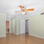 A spacious, empty room with light green walls, light wood flooring, a ceiling fan with wooden blades, and several white doors leading to other areas.