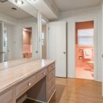 A dressing room with built-in shelves, a large mirror, and a wide counter with drawers. Two open doors reveal a bathroom with light pink tiles and a window. The floor is light wood.