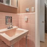 A small bathroom features a pink wall-mounted sink and matching pink tile with a silver faucet, a mirror cabinet above, and a glimpse of a hallway with wood flooring and white doors.