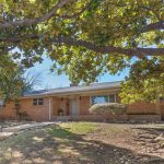 Single-story brick house with a covered porch, surrounded by large trees and landscaping rocks. Sunlight filters through the branches, casting shade on the front yard and walkway.