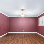 An empty room with reddish walls, a white ceiling, wood flooring, white columns on each side, a central light fixture, and a window with white shutters on the right wall.