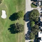 Aerial view of a residential neighborhood next to a golf course with a sand bunker. A marker pin highlights the roof of a house near the street dividing the homes from the golf course. Trees line both sides of the road.