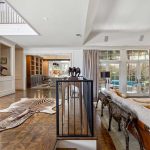 Spacious living room with large windows, patterned wood floor, zebra rug, elegant furniture, decorative sculptures, and an open staircase leading to an upper level. Light fills the room from tall glass doors and windows.