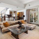 Spacious living room with a beige sectional sofa, glass coffee table, tan armchair, bookshelves, and large windows opening to a patio. A staircase and dark wood double doors are visible in the background.