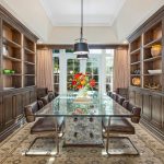 A dining room with a glass table, brown leather chairs, wooden shelves and cabinets, a floral centerpiece, and a large window letting in natural light. Shelves hold various decorative items.