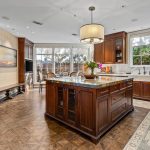 Spacious kitchen with dark wood cabinets, large marble island, pendant light, two rugs on patterned wooden floor, and large windows letting in natural light. Artwork and a statue are visible on the left wall.