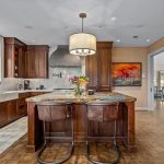 A spacious kitchen with dark wood cabinets, a marble island with two brown chairs, stainless steel appliances, a modern light fixture, flowers on the island, and large windows letting in natural light.