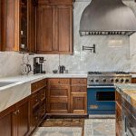 A kitchen with wooden cabinets, a marble countertop and backsplash, a large metal range hood, a blue stove, a built-in sink, and a coffee maker under a window with greenery outside.